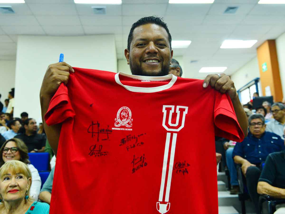 Pedro Lemus sostiene y muestra sonriente la camiseta de la UES autografiada por «Pipo» Rodríguez y otros futbolistas.