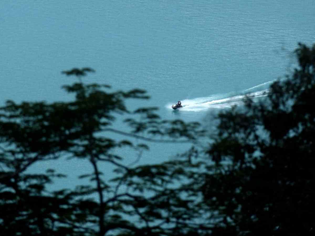 Vista lejana de una moto acuática navegando a gran velocidad por el agua turquesa del Lago de Coatepeque, enmarcada por vegetación.