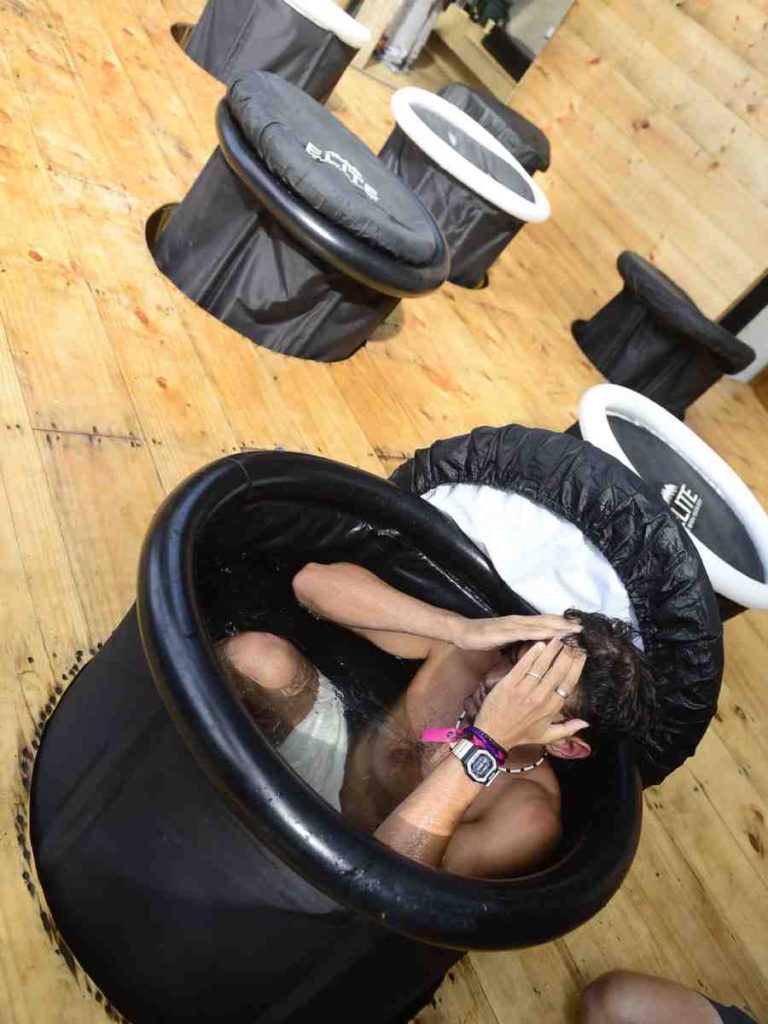 Atleta sumergido en una tina portátil de hieloterapia instalada sobre una plataforma de madera en la zona de recuperación.